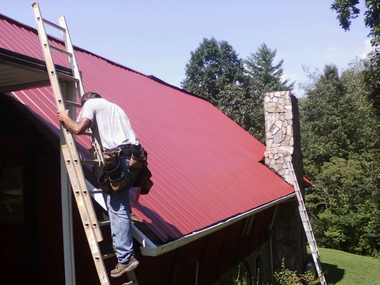 Roofer on a ladder beside a red metal roof and stone chimney — Soffit & Fascia Repair by Roofing Brothers in Salix